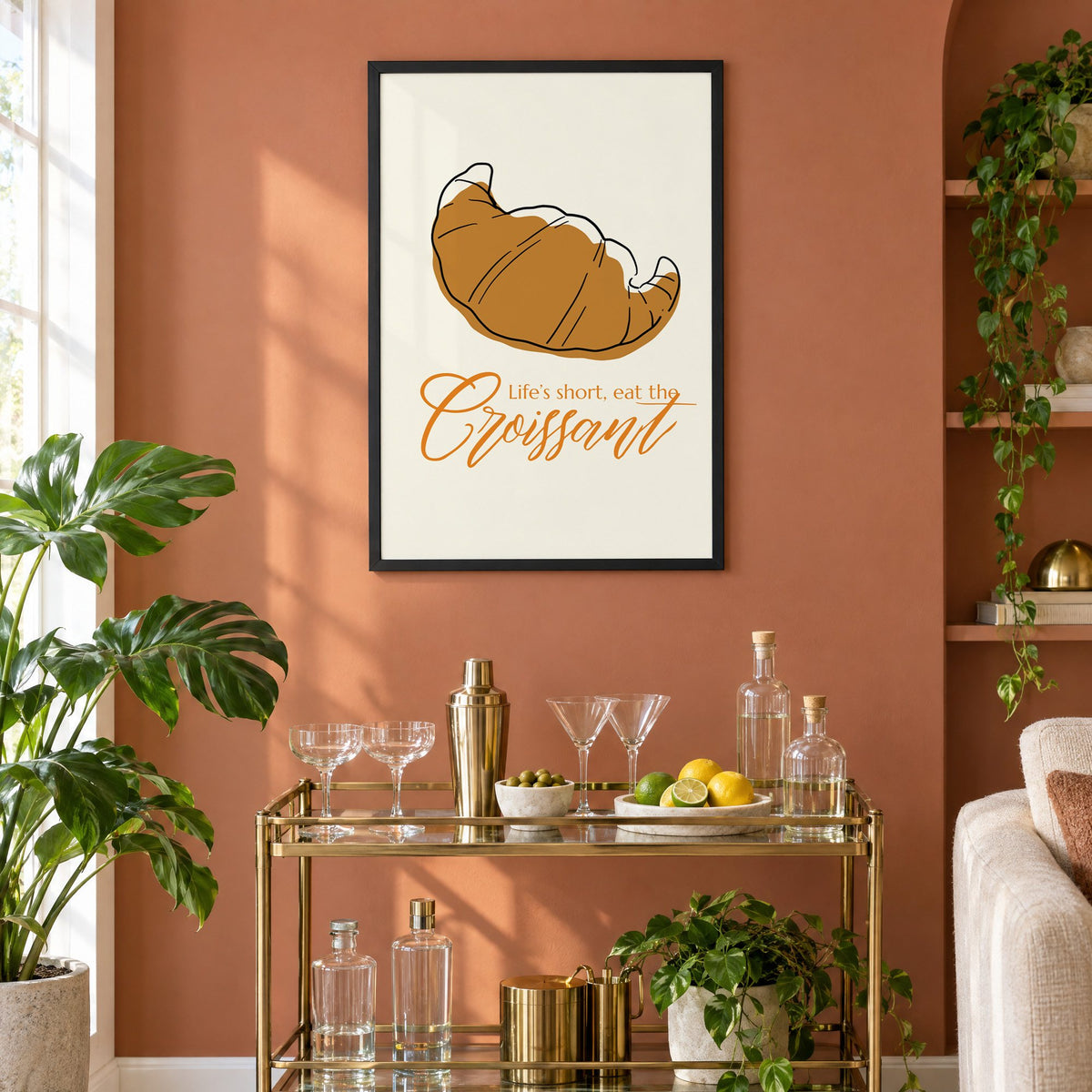 Life's short, eat the Croissant print in a black frame on a terracotta wall above a brass bar cart with coupes and citrus