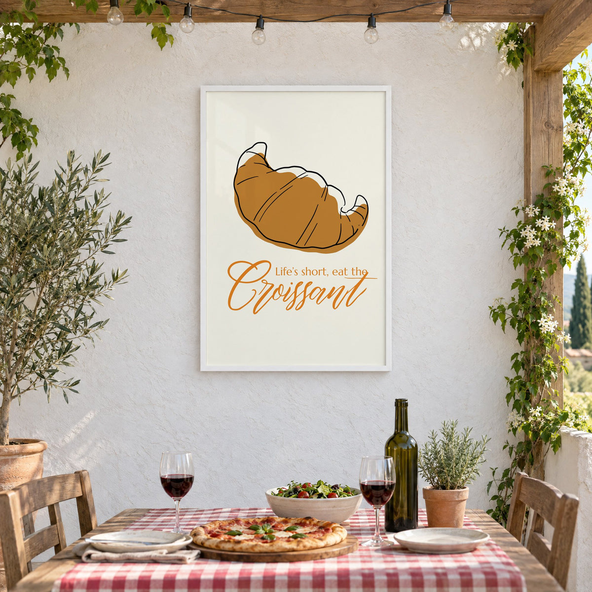 Life's short, eat the Croissant print in a white frame on a stucco wall above a checked-cloth table with pizza and red wine
