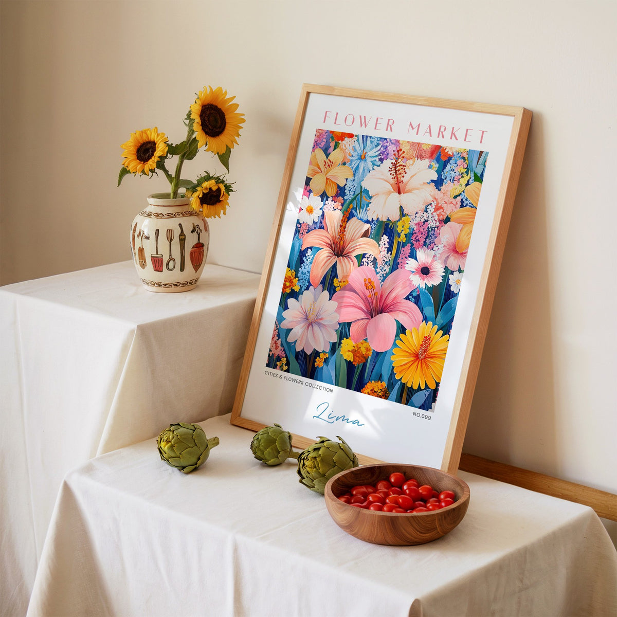 Lima flower market print in an oak frame leaning on a linen-draped table beside a vase of sunflowers
