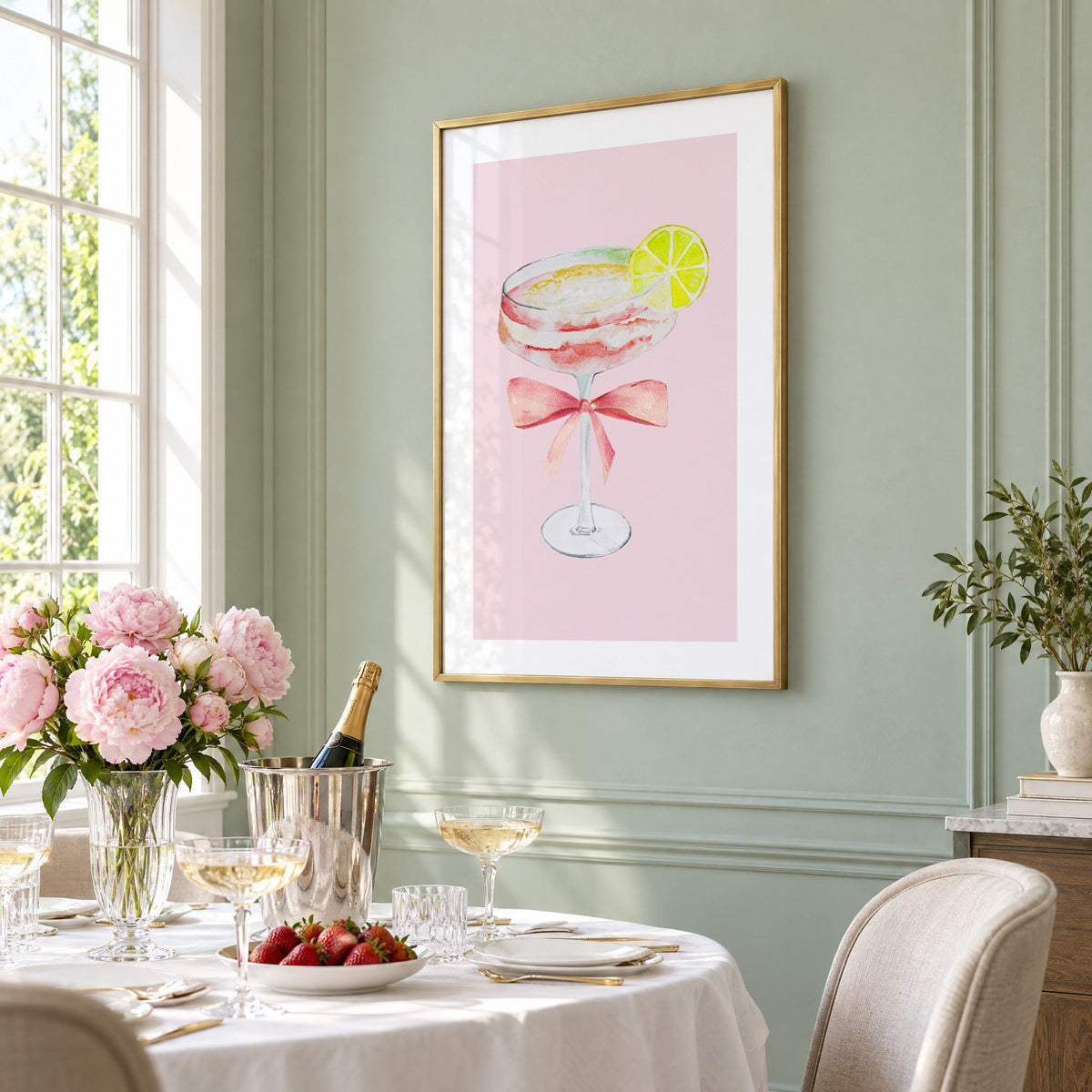 lime coupe cocktail pink bow watercolor print in a brass frame on a sage wall above a brunch table with champagne and peonies, seen at an angle
