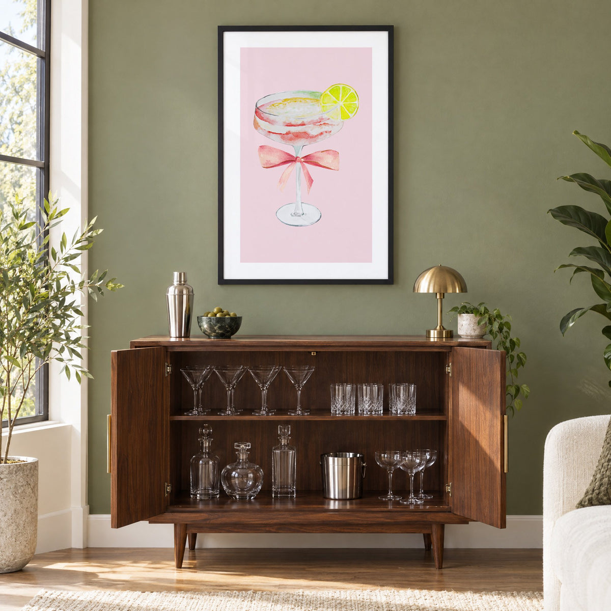 lime coupe cocktail pink bow watercolor print in a black frame on an olive green wall above a walnut bar cabinet with glassware