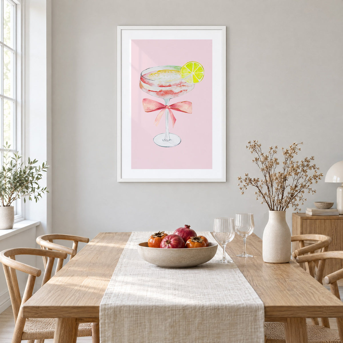 lime coupe cocktail pink bow watercolor print in a white frame above a light oak dining table with a linen runner and fruit bowl