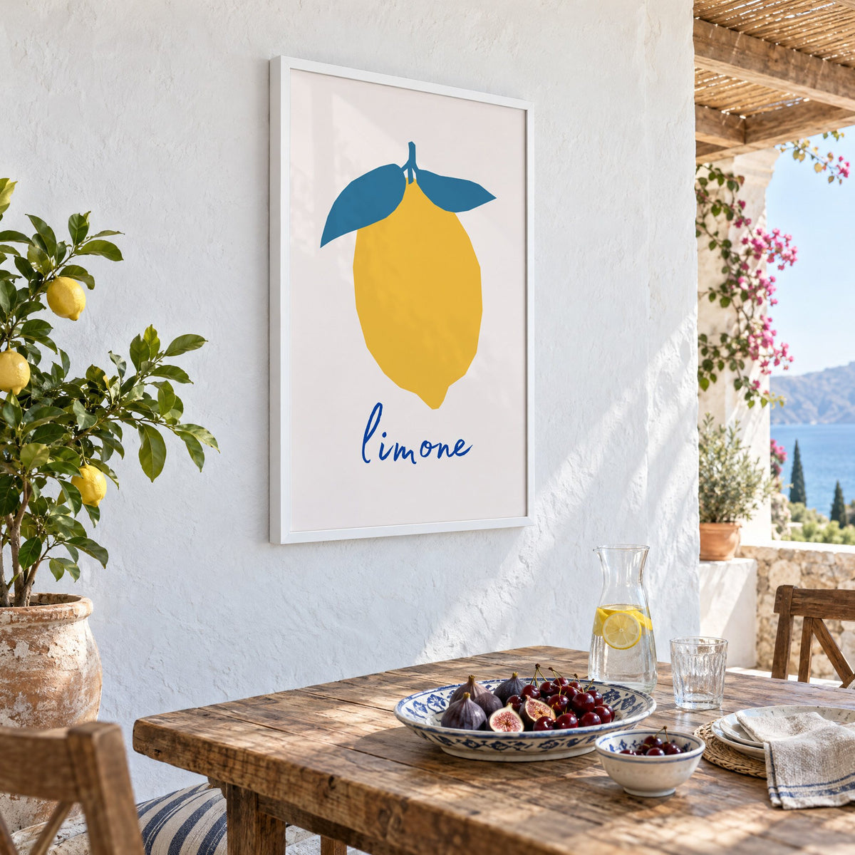 limone yellow cut-paper lemon print in a white frame on a whitewashed terrace wall above a lunch table with figs and cherries, seen at an angle