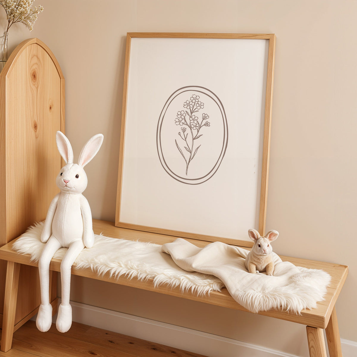 Line art flower sprig in a double oval nursery print in an oak frame on a sheepskin bench beside a white plush bunny