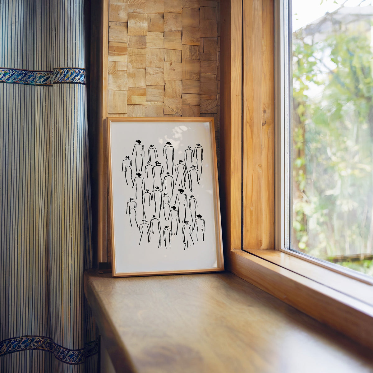 Crowd of hat-wearing figures line art print in a light wood frame leaning on a wooden window sill by a garden view