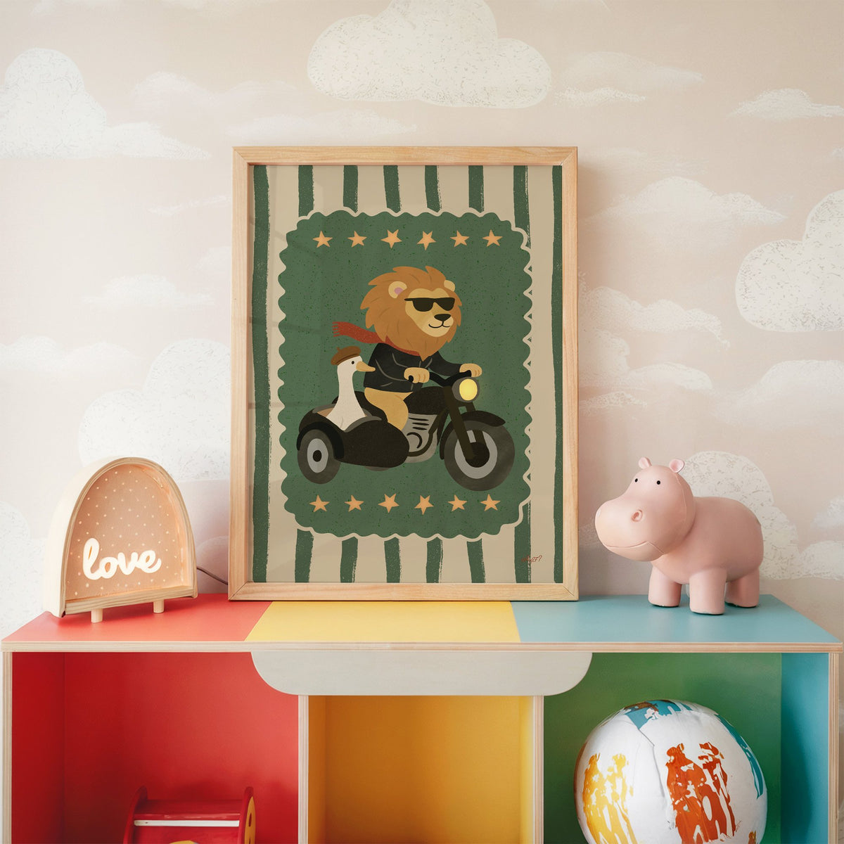 Lion on motorcycle with goose sidecar nursery print in an oak frame on a rainbow toy shelf against cloud wallpaper