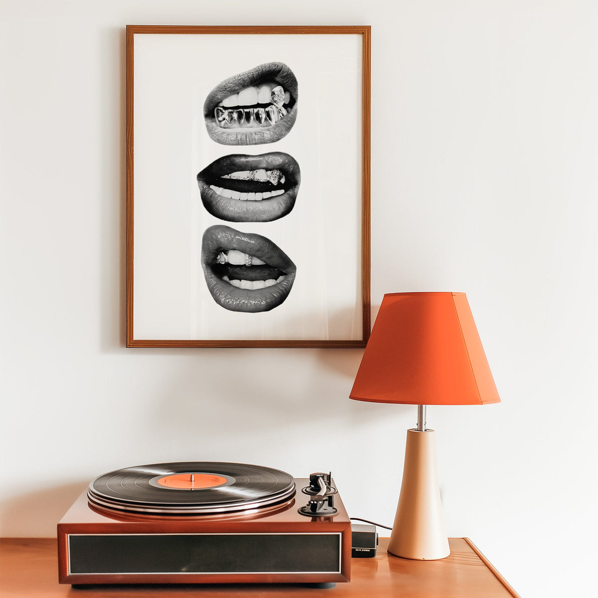 Three lips with jeweled metal grills print in a walnut frame above a record player and orange lamp