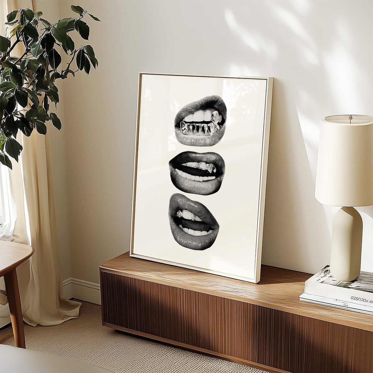 Three lips with jeweled metal grills print in a white frame on a walnut sideboard beside a ceramic lamp