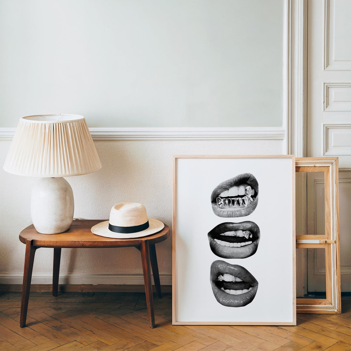 Three lips with jeweled metal grills print in a pine frame on a wood floor beside a lamp and straw hat