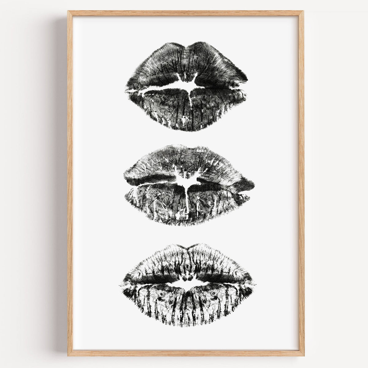 Three lipstick kiss marks stacked print in a light oak frame on a plain white wall