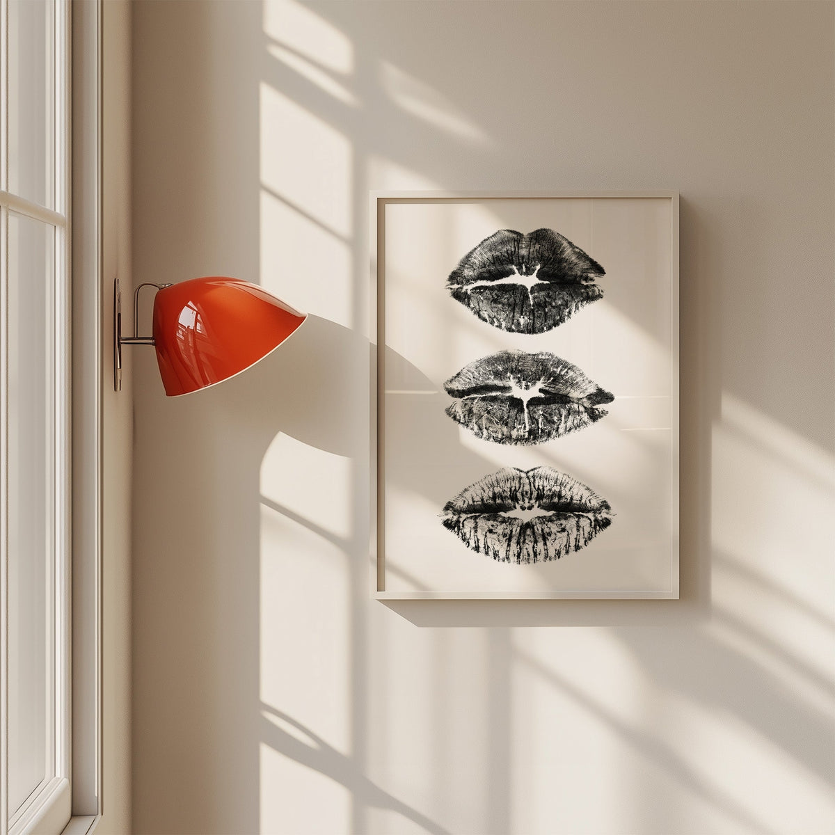 Three lipstick kiss marks stacked print in a white frame on a sunlit wall beside a red sconce