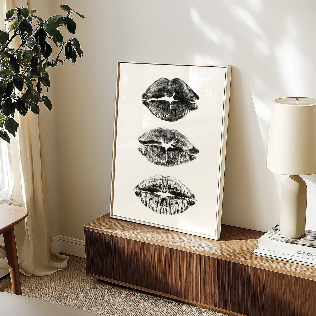 Three lipstick kiss marks stacked print in a white frame on a walnut sideboard beside a ceramic lamp