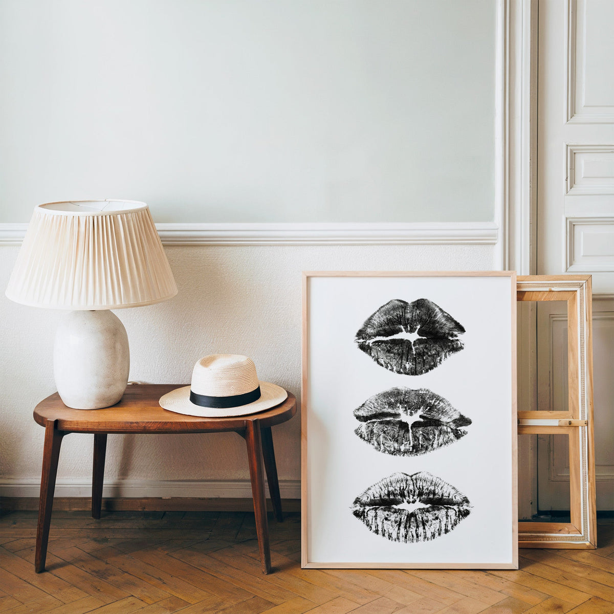 Three lipstick kiss marks stacked print in a pine frame on a wood floor beside a lamp and straw hat