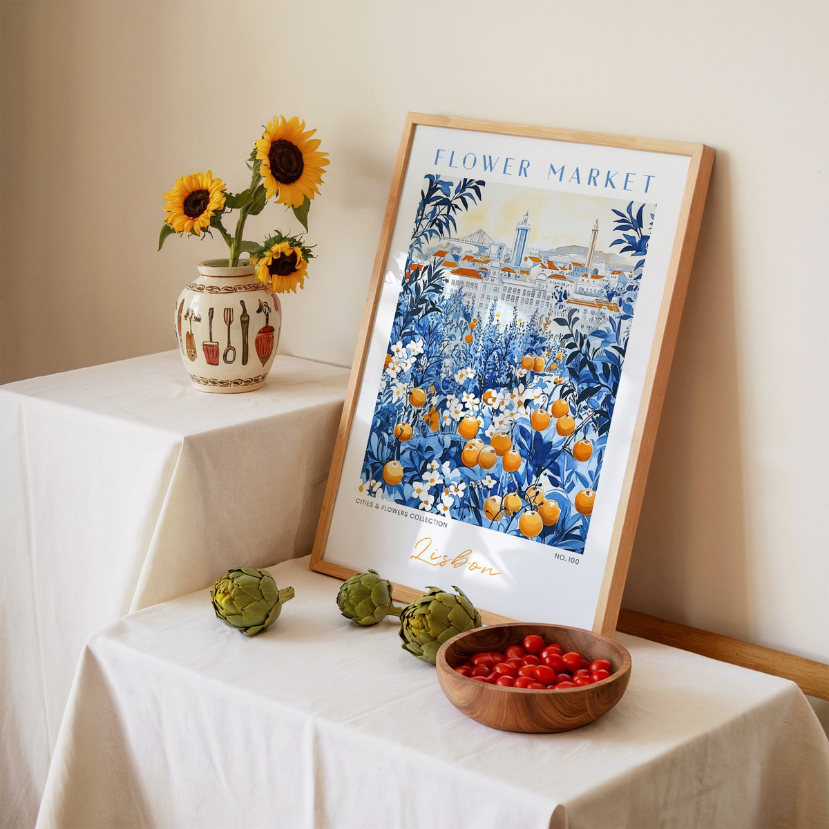 Lisbon flower market print in an oak frame leaning on a linen-draped table beside a vase of sunflowers