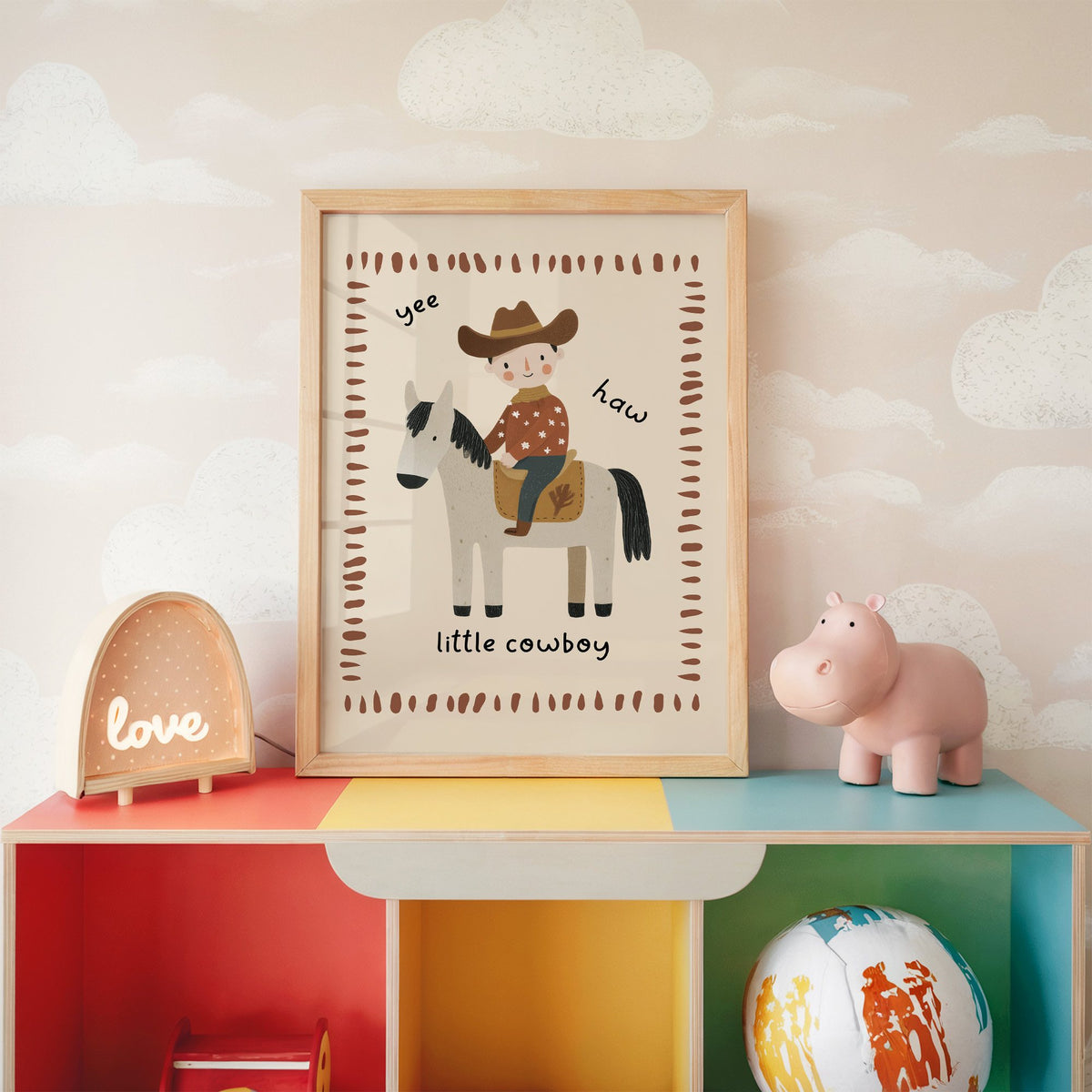 Little cowboy on horse yee haw nursery print in an oak frame on a rainbow toy shelf against cloud wallpaper