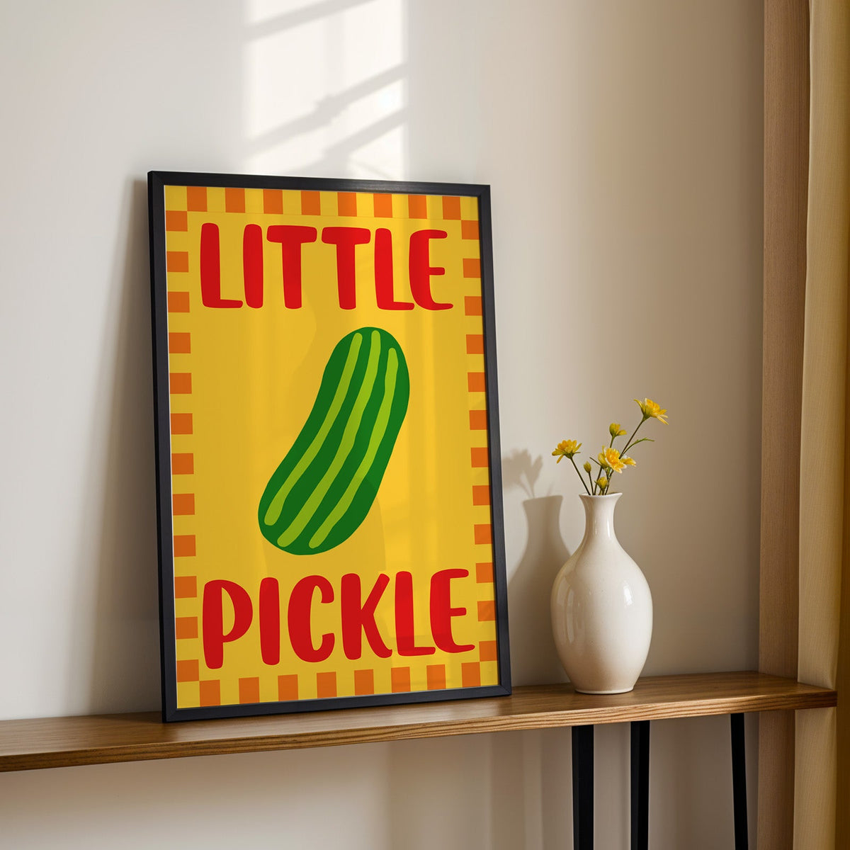 Little pickle print in a black frame on a console table with yellow daisies