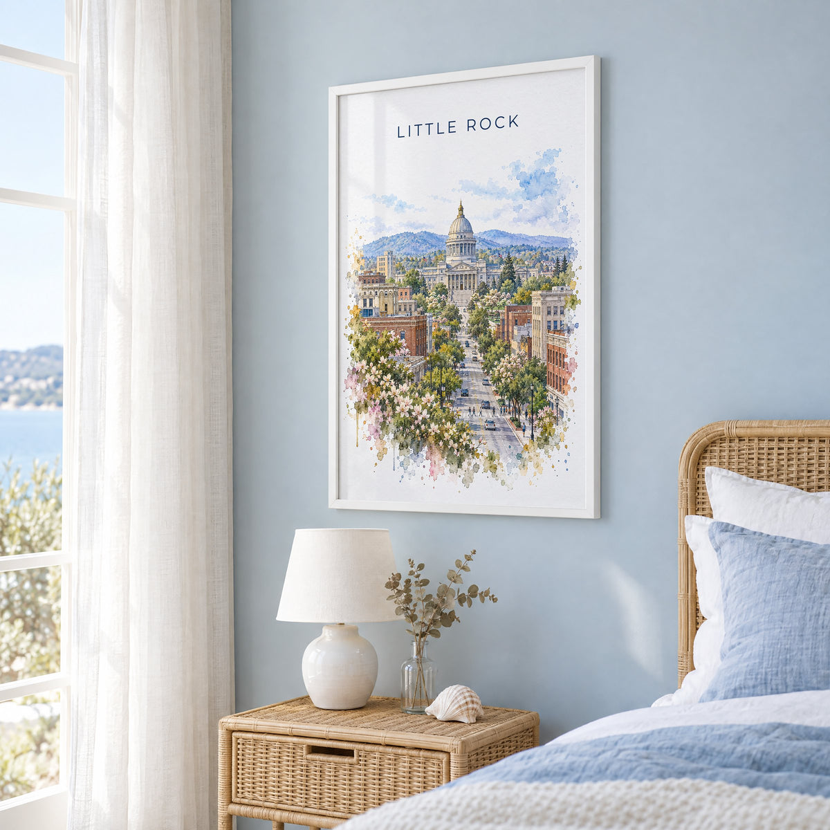 Little Rock watercolor print in a white frame on a pale blue wall beside a rattan bed in a coastal bedroom
