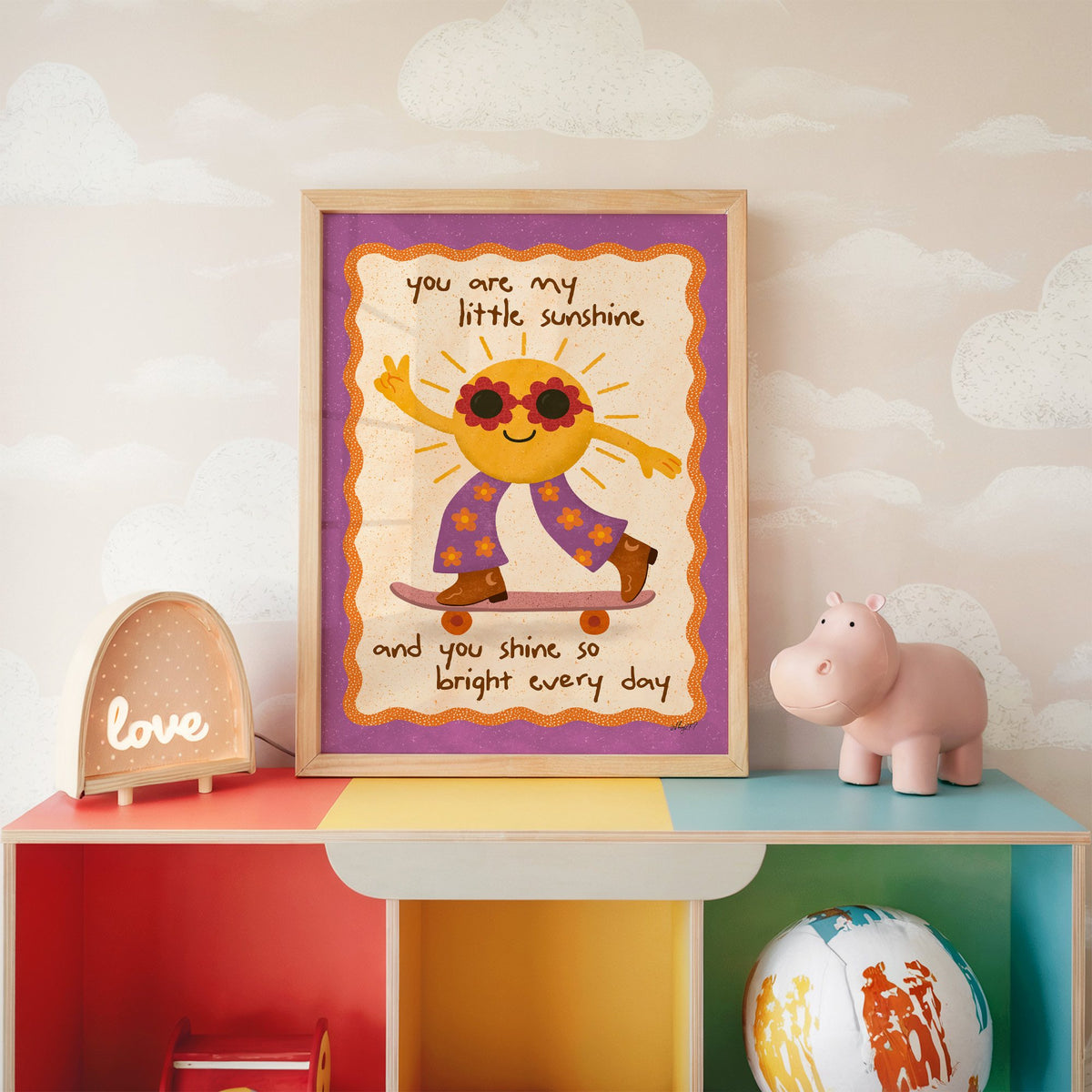 Skateboarding sun little sunshine nursery print in an oak frame on a rainbow toy shelf against cloud wallpaper