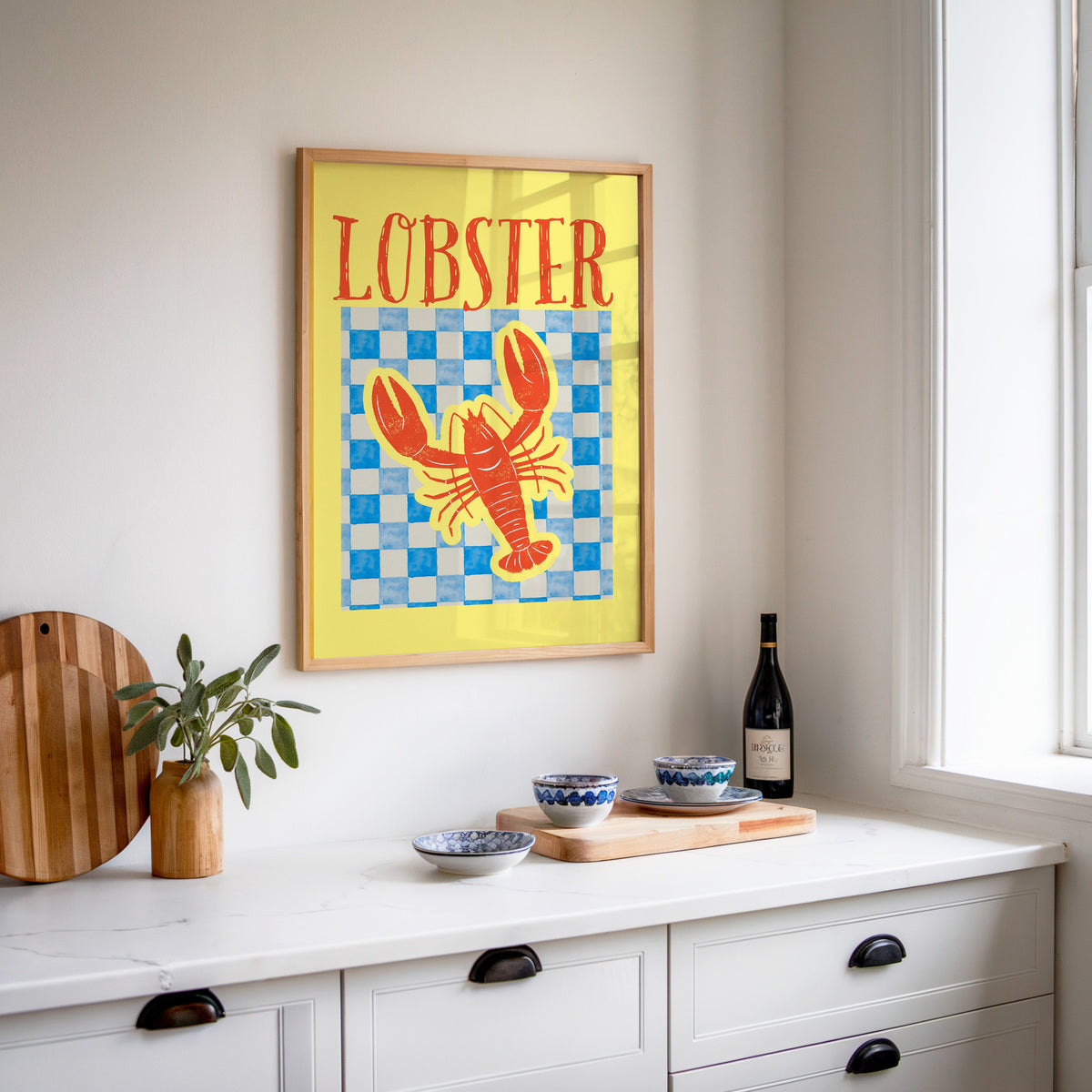 Lobster blue check print in an oak frame above a white sideboard with a bottle of wine