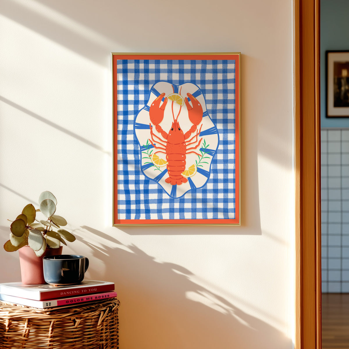 Lobster gingham print shown in a gold frame above a basket of books and a pilea plant