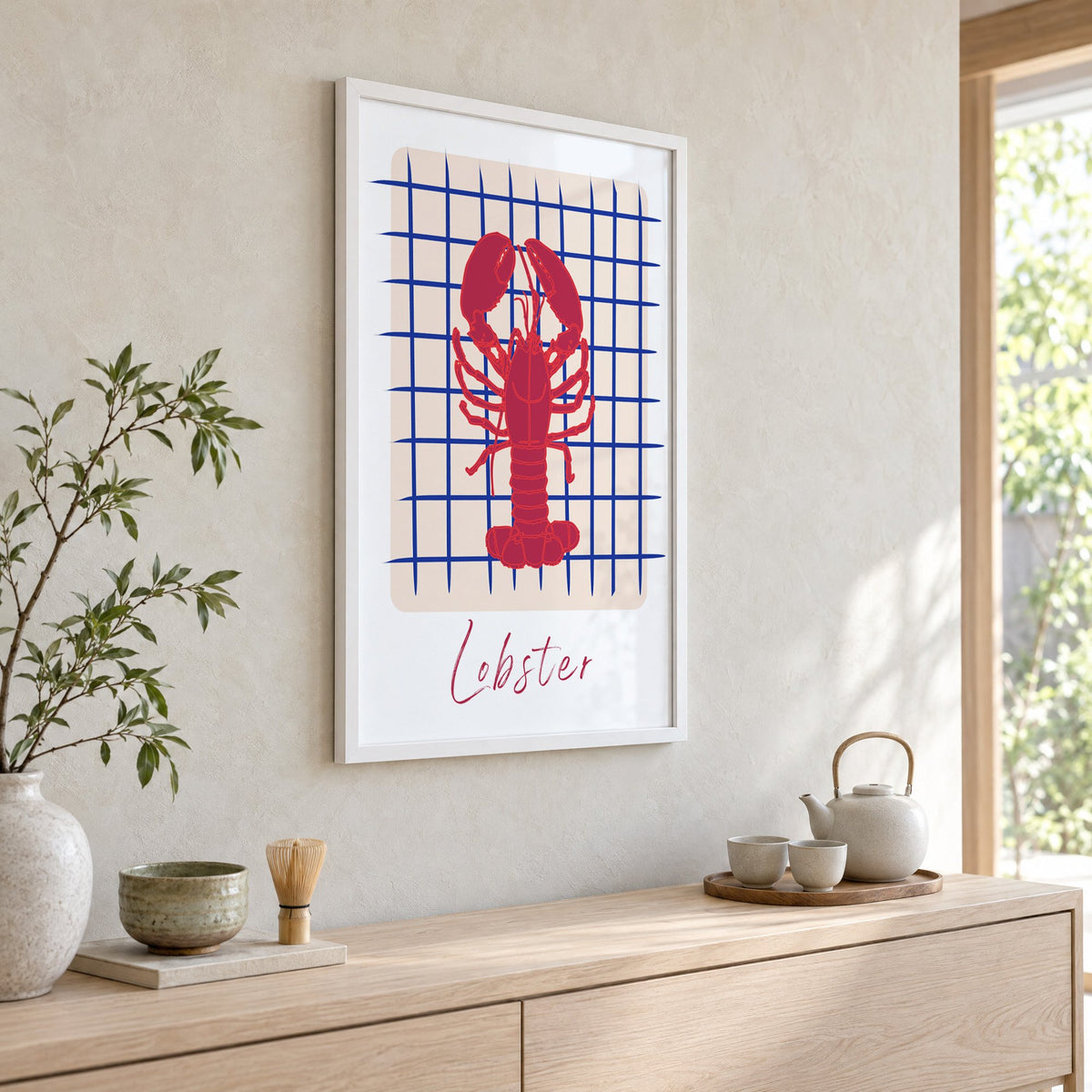Lobster on cobalt grid print in a white frame above a pale wood console with a matcha tea set