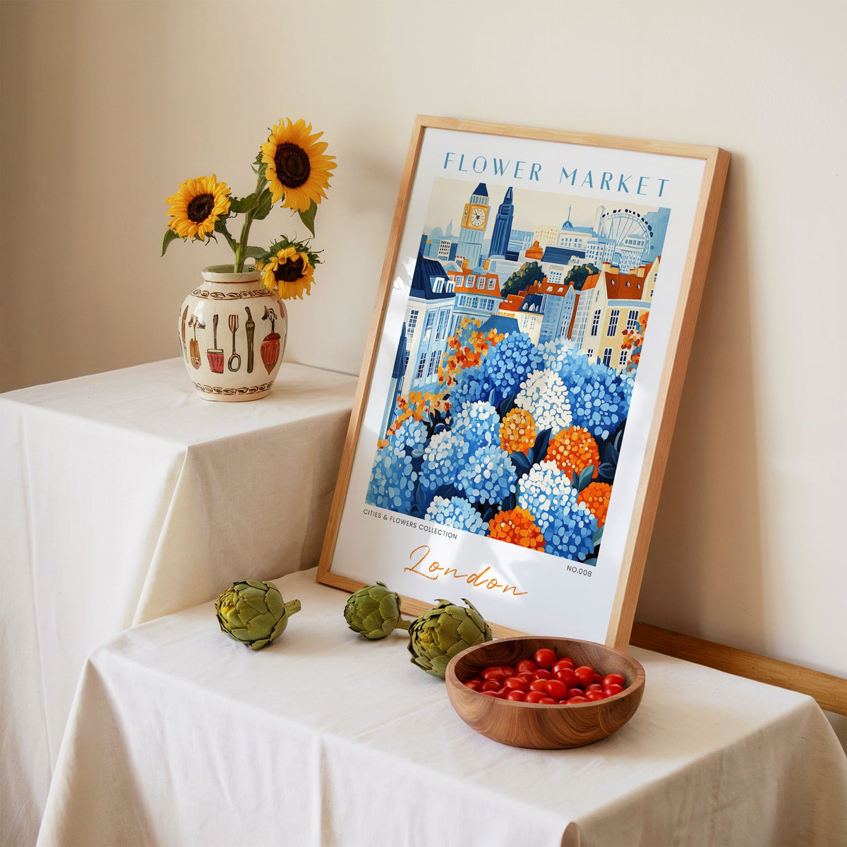 London flower market print in an oak frame leaning on a linen-draped table beside a vase of sunflowers