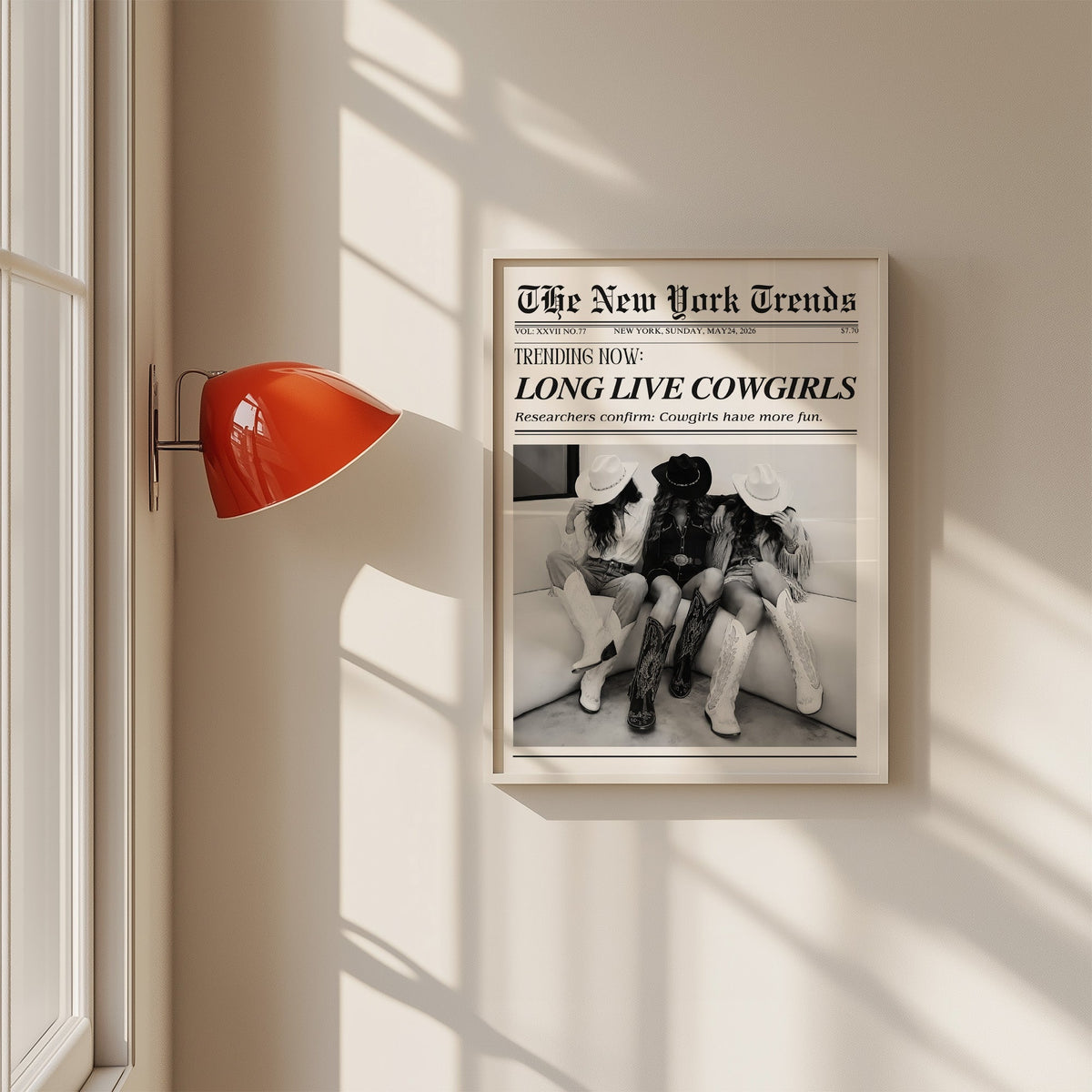 Three cowgirls on white sofa mock newspaper print in a white frame on a sunlit wall beside a red sconce