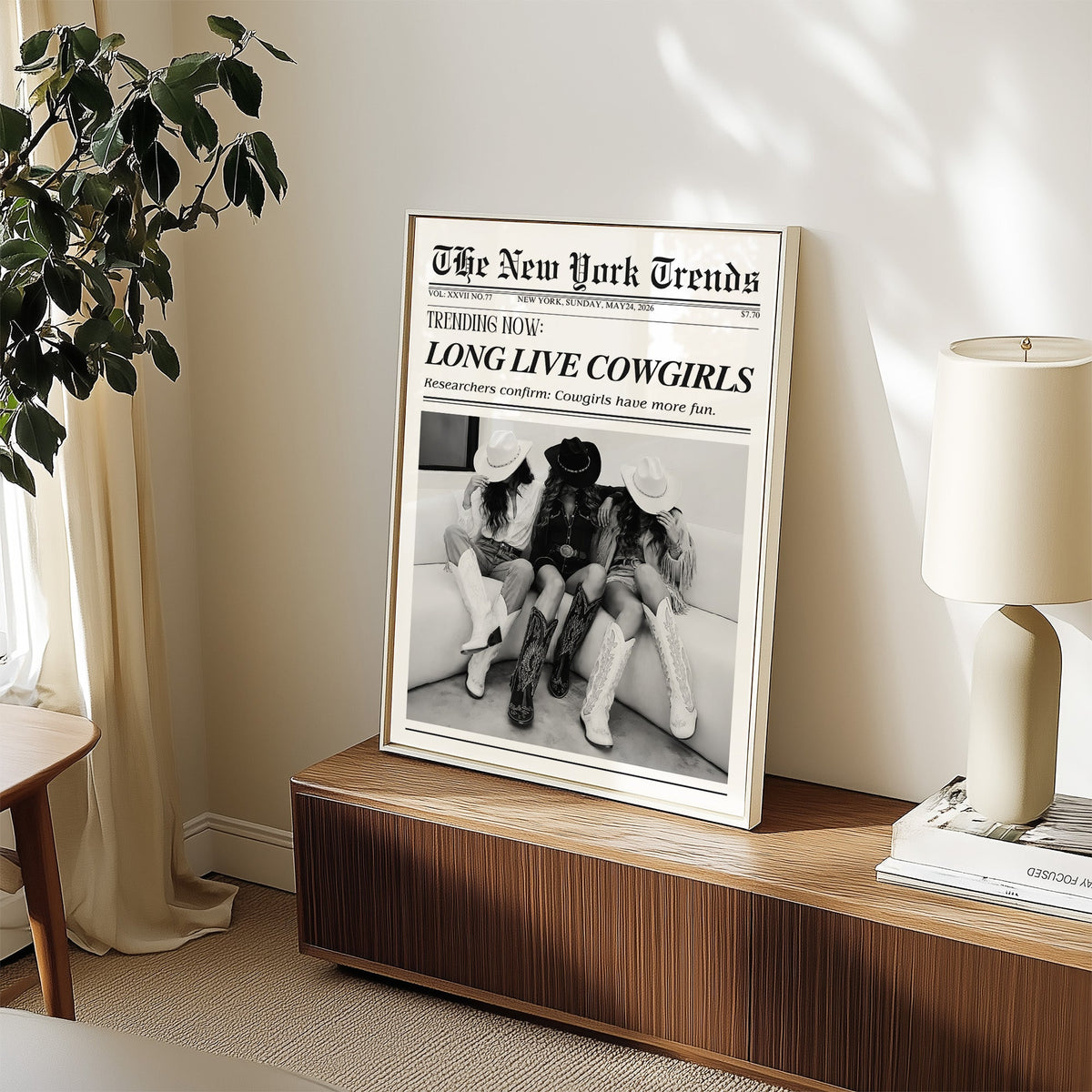 Three cowgirls on white sofa mock newspaper print in a white frame on a walnut sideboard beside a ceramic lamp