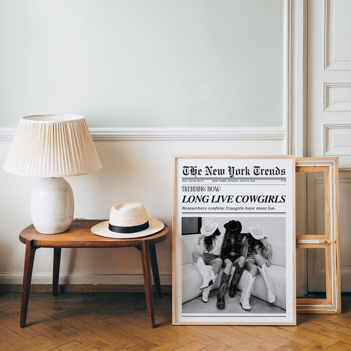 Three cowgirls on white sofa mock newspaper print in a pine frame on a wood floor beside a lamp and straw hat