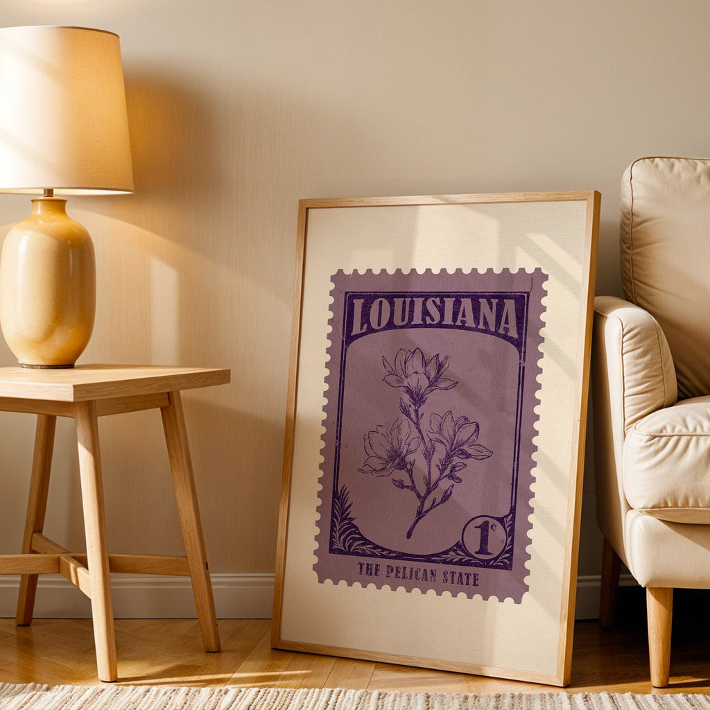 Louisiana stamp print in a wood frame resting on the floor beside a beige armchair and a yellow lamp