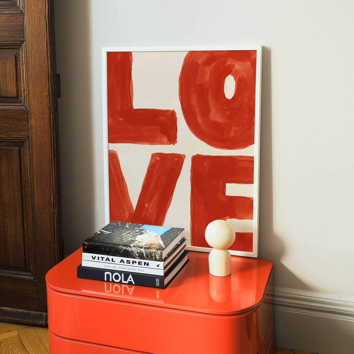 Red watercolor LOVE print in a white frame on a glossy orange cabinet with books