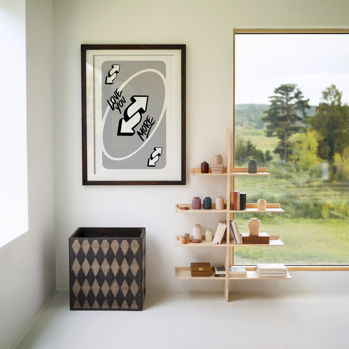 Love you more reverse arrows card print in a walnut frame with a white mat beside wooden shelves and a garden window