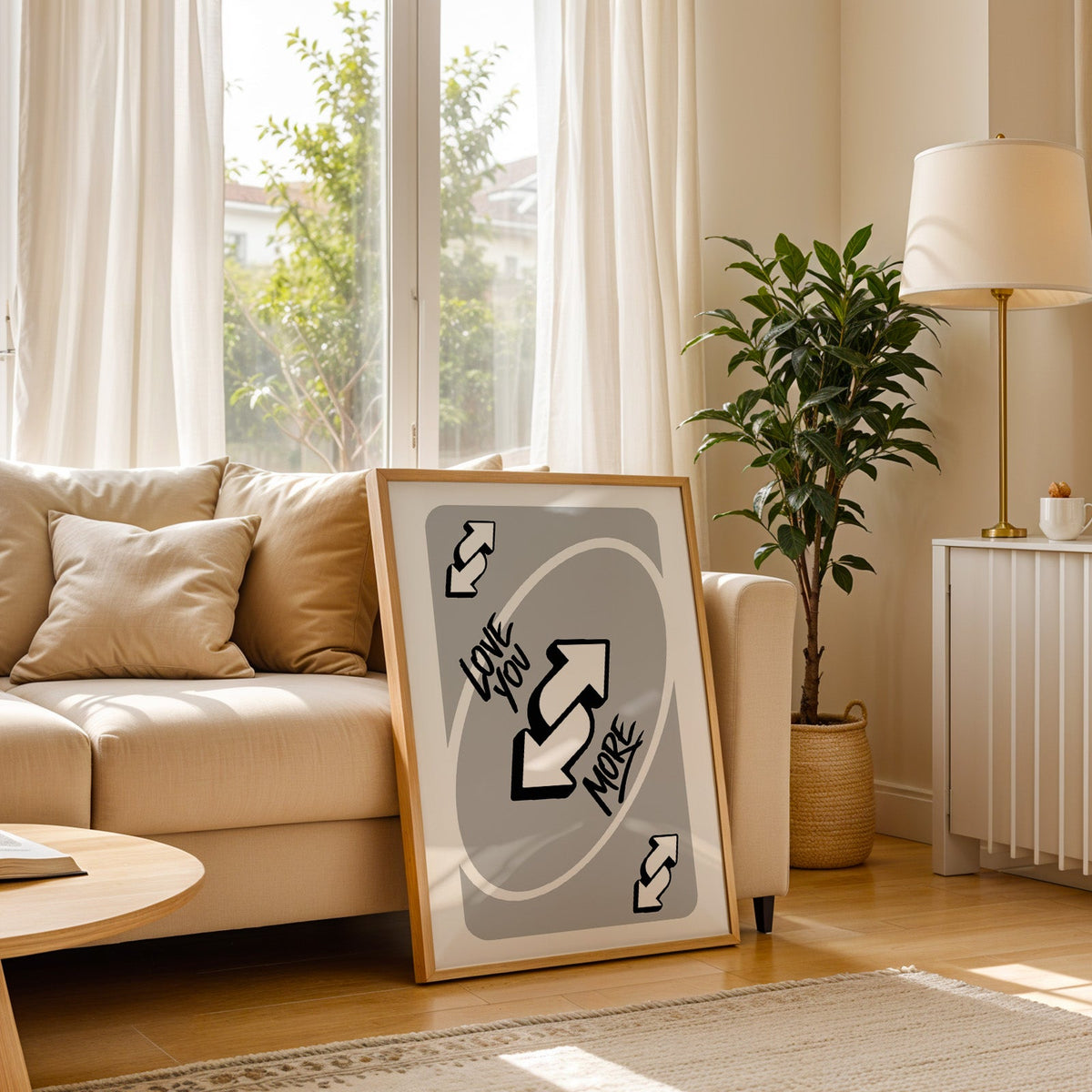 Love you more reverse arrows card print in a light oak frame leaning on a cream sofa beside a floor lamp
