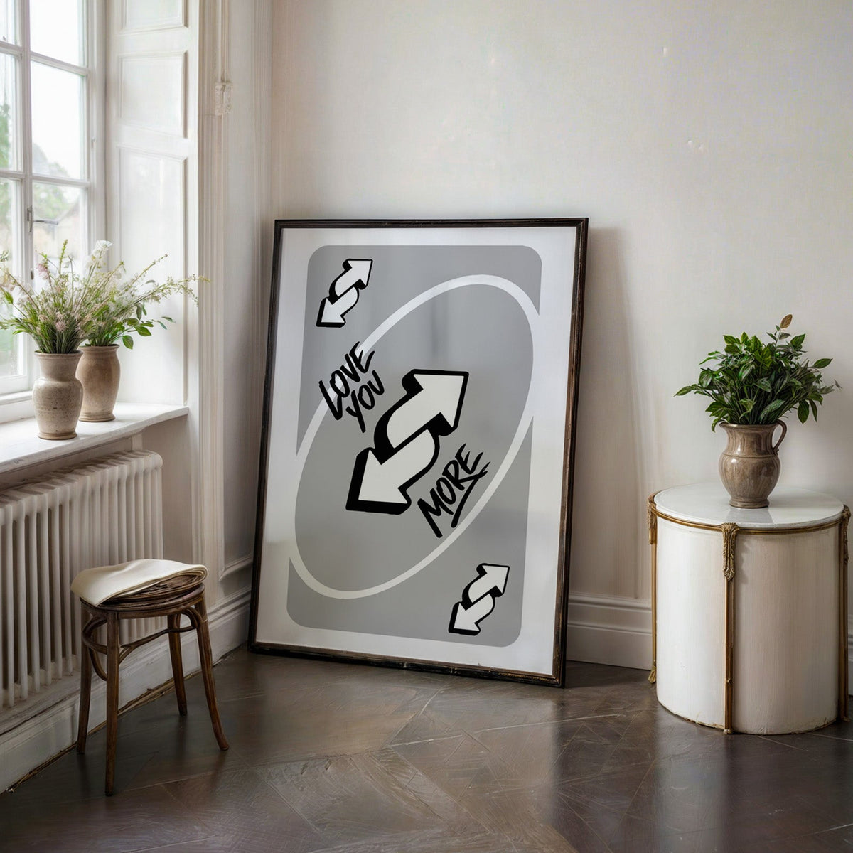Love you more reverse arrows card print in a black frame on a dark stone floor beside a white side table