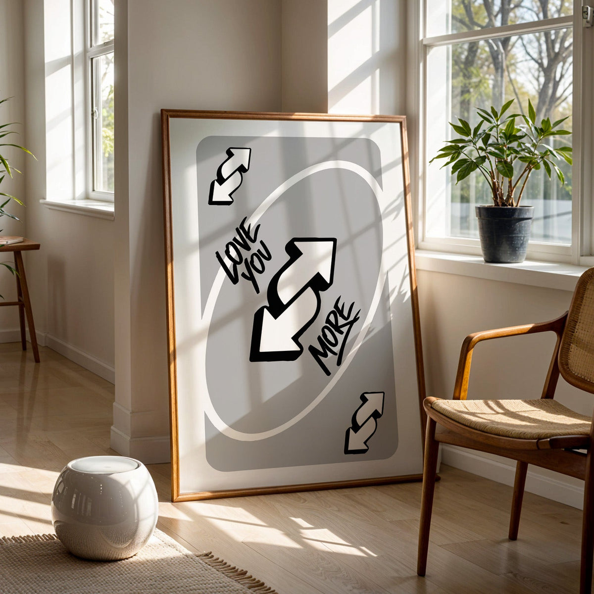 Love you more reverse arrows card print in a walnut frame on a wood floor beside a cane chair and sunny window