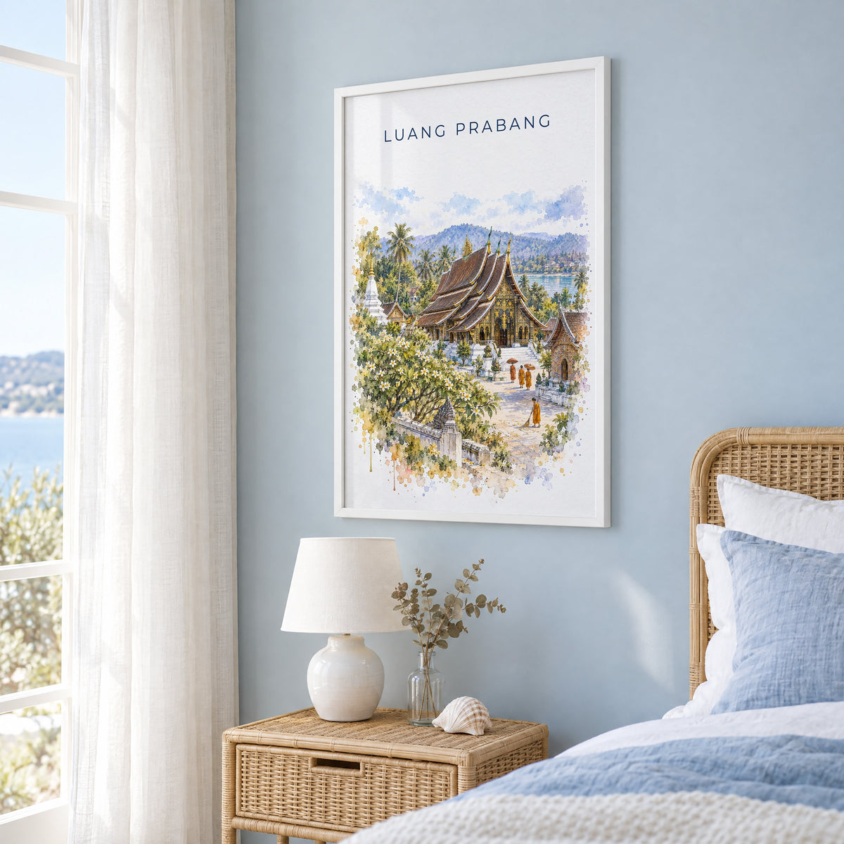 Luang Prabang watercolor print in a white frame on a pale blue wall beside a rattan bed in a coastal bedroom