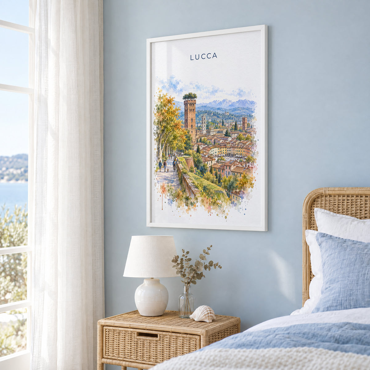 Lucca watercolor print in a white frame on a pale blue wall beside a rattan bed in a coastal bedroom