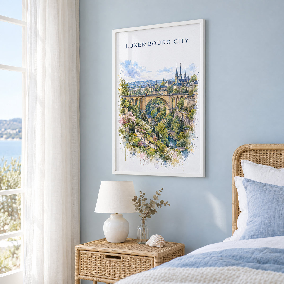 Luxembourg City watercolor print in a white frame on a pale blue wall beside a rattan bed in a coastal bedroom