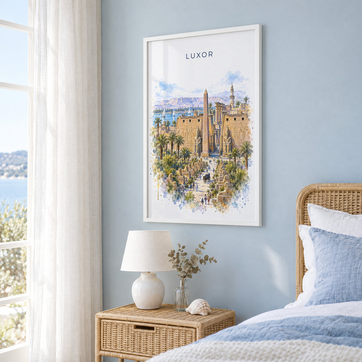 Luxor watercolor print in a white frame on a pale blue wall beside a rattan bed in a coastal bedroom