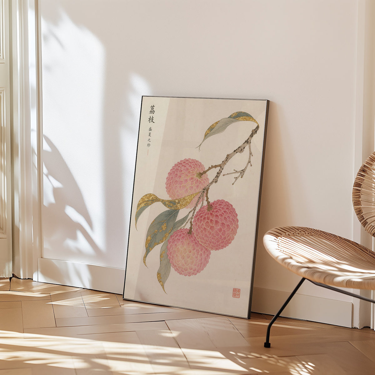 lychee branch with gold-toned leaves print in a black frame leaning on a herringbone floor beside a woven rattan chair