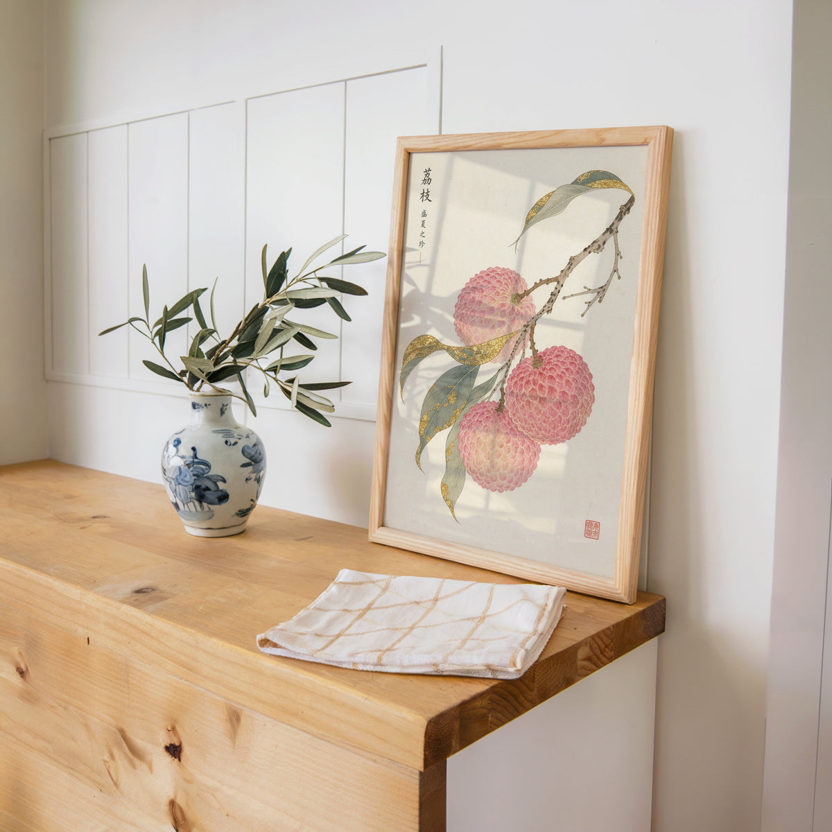 lychee branch with gold-toned leaves print in a pine frame on a wooden counter beside a blue and white porcelain vase