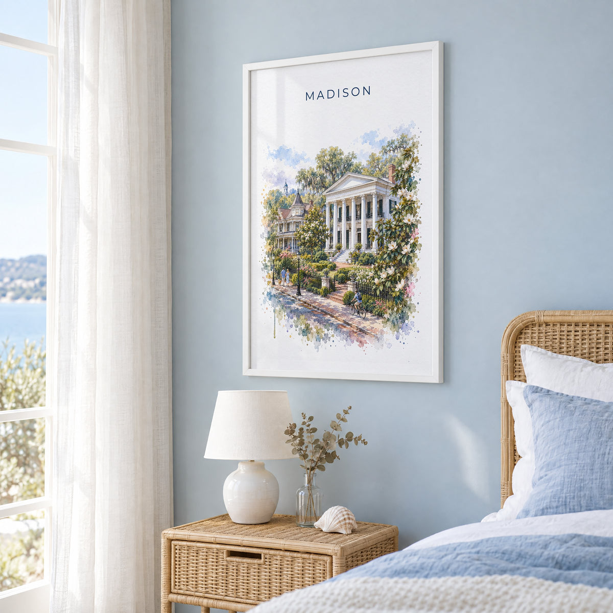 Madison, Georgia watercolor print in a white frame on a pale blue wall beside a rattan bed in a coastal bedroom