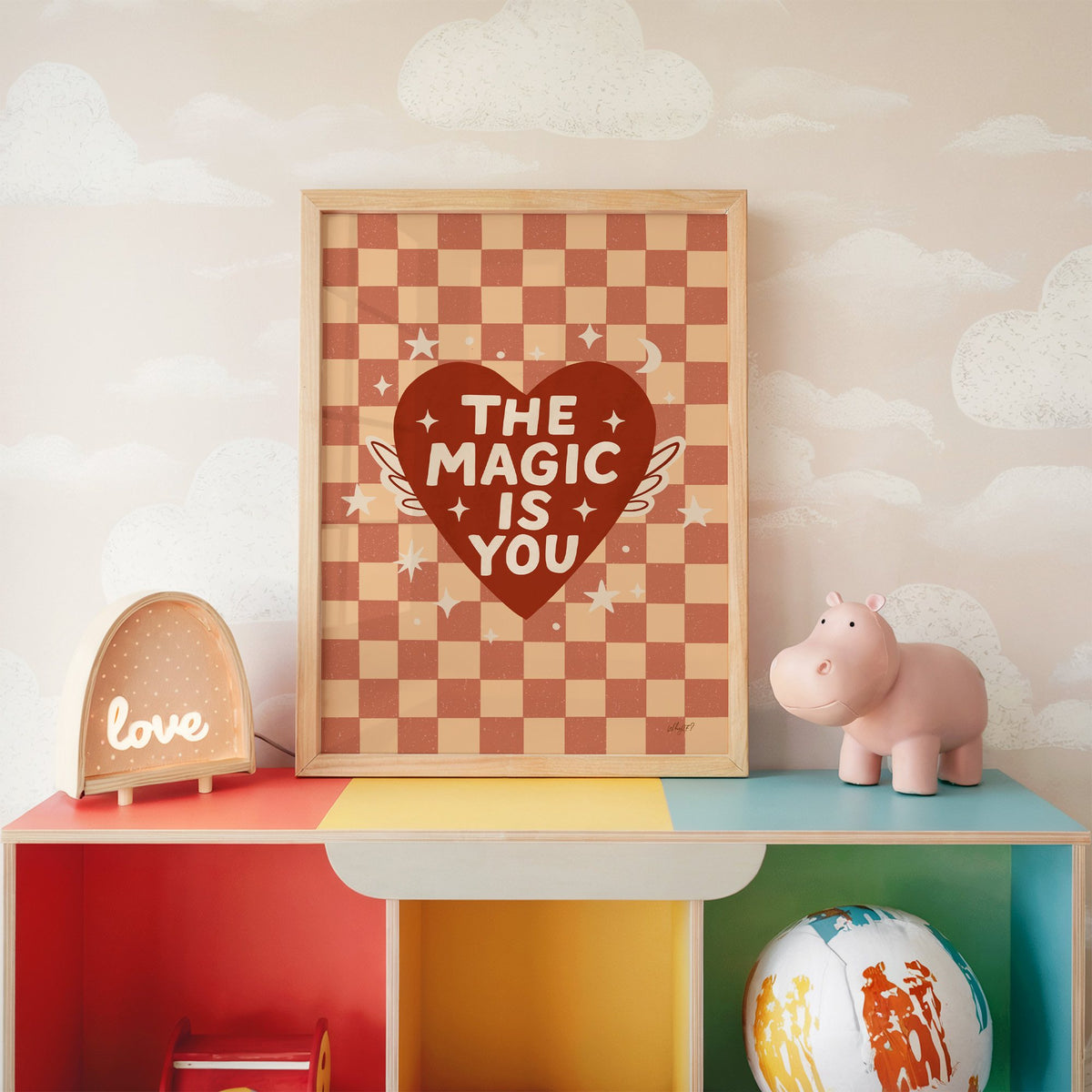 The magic is you winged heart nursery print in an oak frame on a rainbow toy shelf against cloud wallpaper