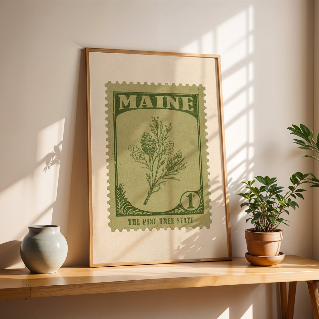 Maine White Pine Cone and Tassel stamp print in a light oak frame leaning on a wooden table beside a vase