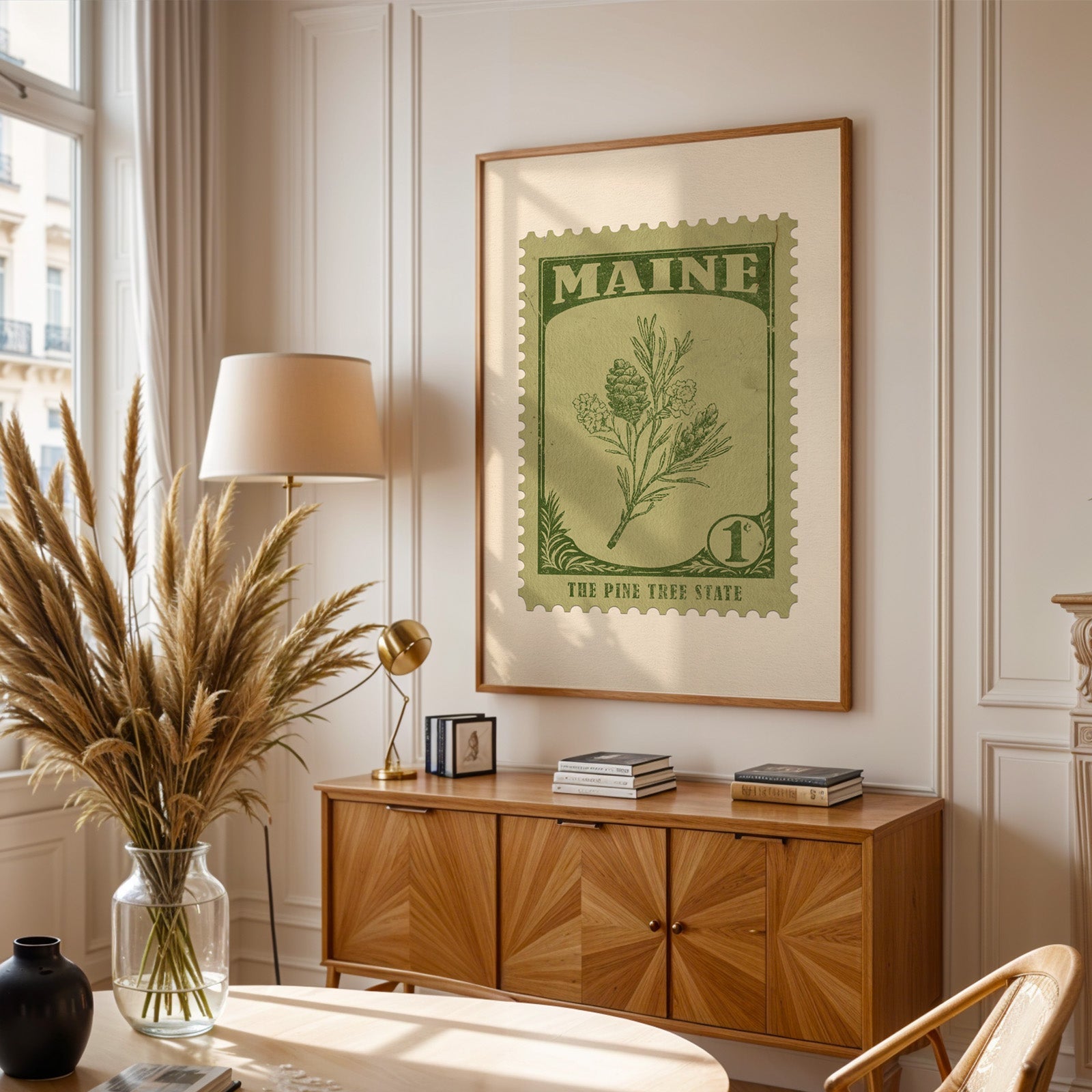 Maine stamp print framed above a wooden sideboard, pampas grass in a glass vase and a floor lamp