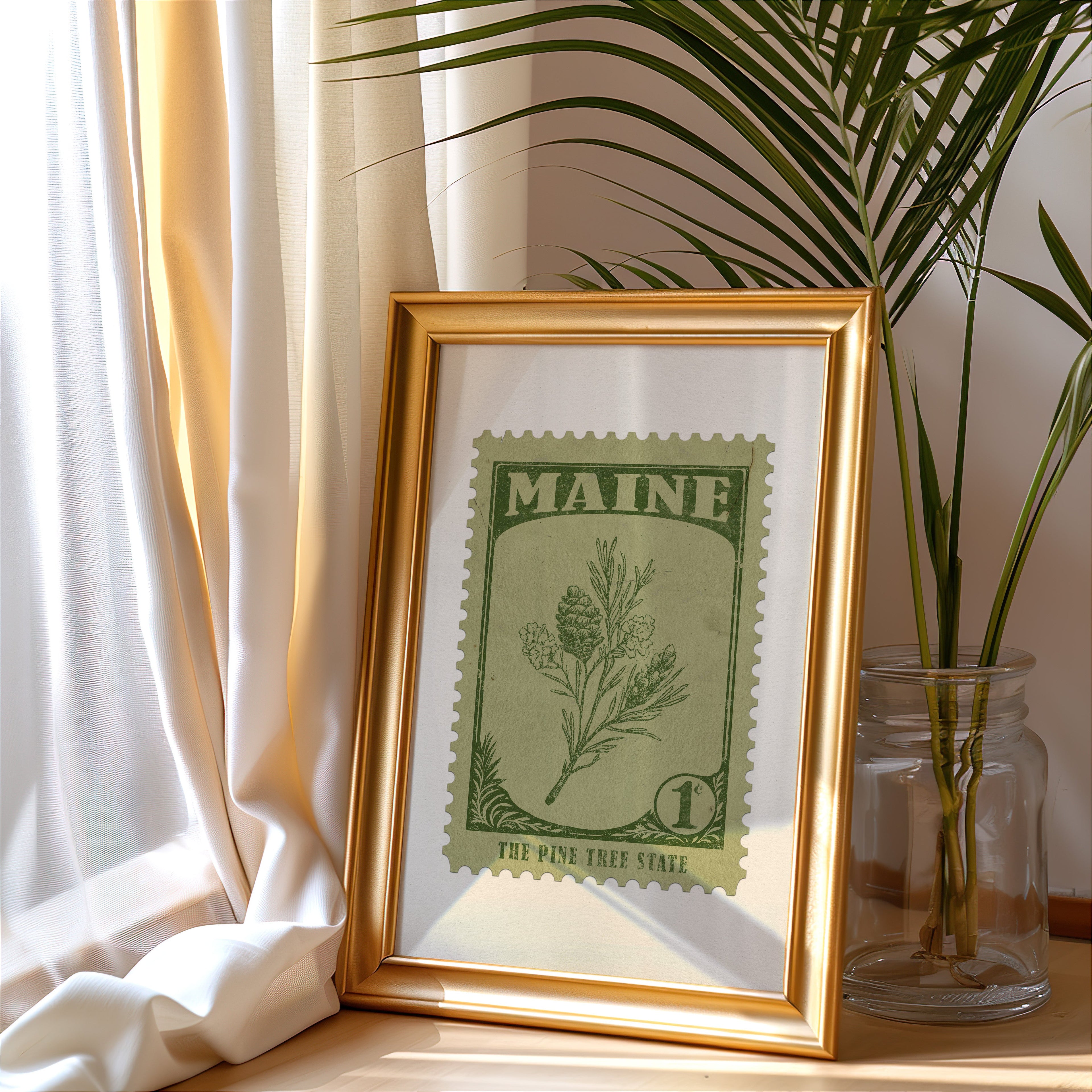 Maine stamp print in a gold frame leaning by a cream curtain with a palm in a glass jar