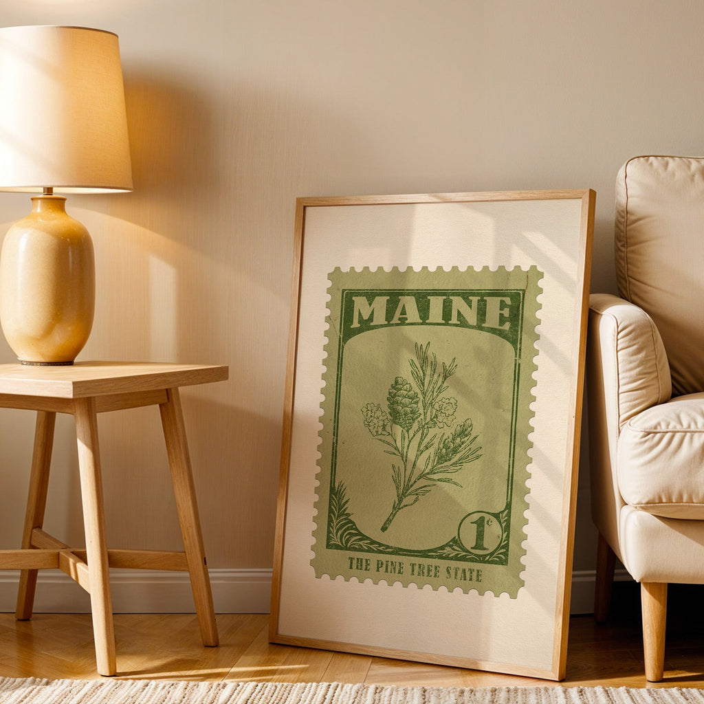 Maine stamp print in a wood frame resting on the floor beside a beige armchair and a yellow lamp
