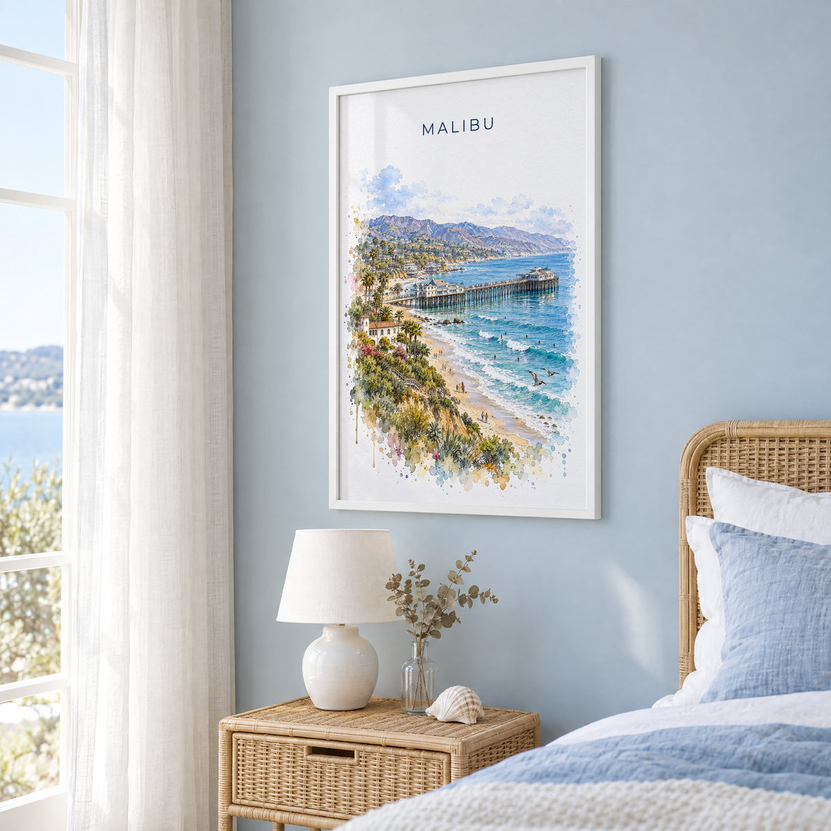 Malibu watercolor print in a white frame on a pale blue wall beside a rattan bed in a coastal bedroom