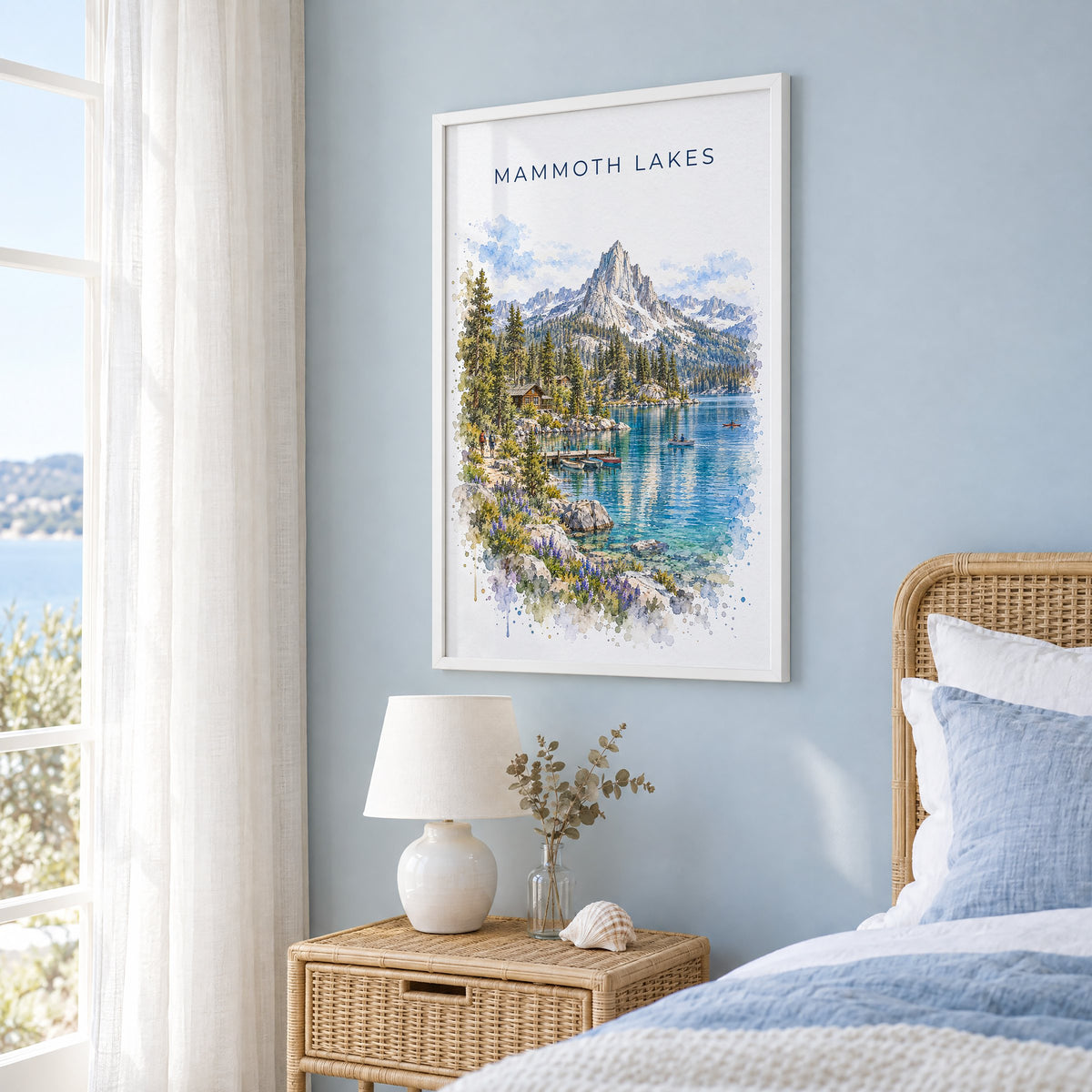 Mammoth Lakes watercolor print in a white frame on a pale blue wall beside a rattan bed in a coastal bedroom