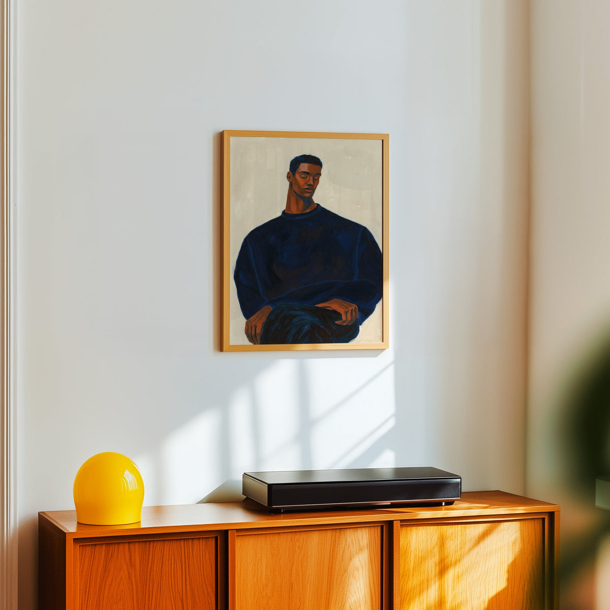 Seated man portrait print in a light oak frame above a teak sideboard with a yellow lamp